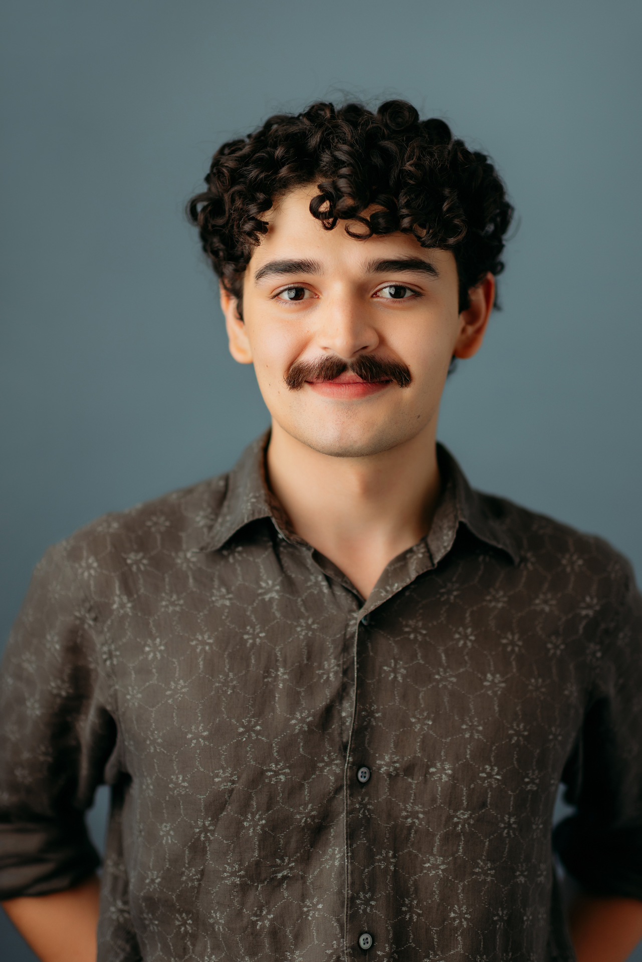 Professional actor headshot of young man with curly hair wearing gray shirt on light blue gray backdrop