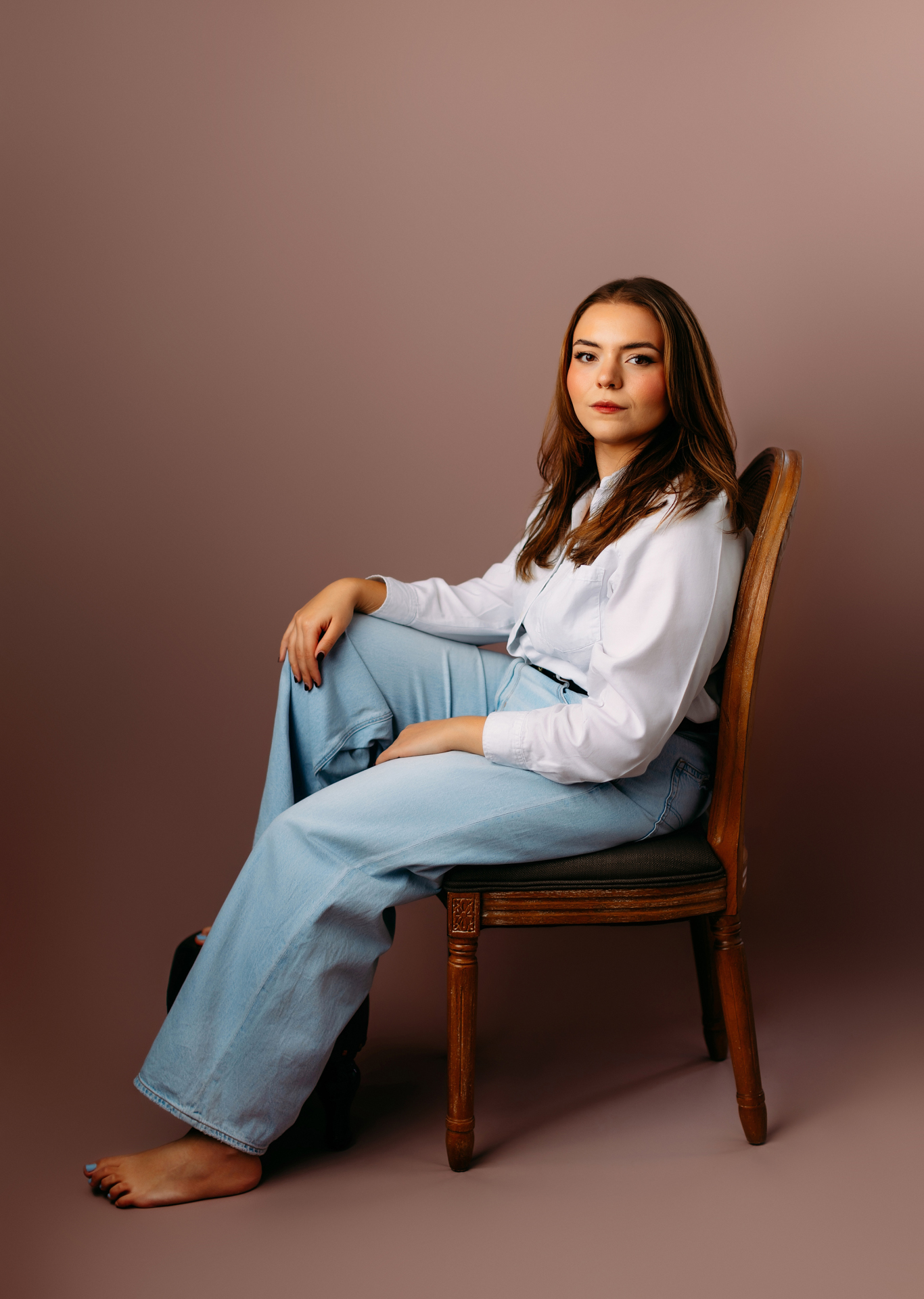 Professional theatre headshot of woman in white pants and light blue jeans sitting profile on wooden chair